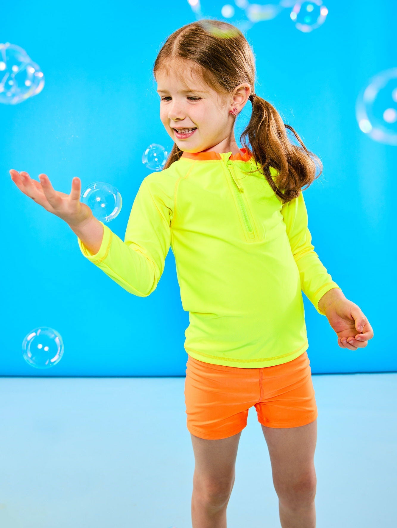 Child plays with bubbles while wearing a neon yellow and neon orange rash guard that features a zipper for easy on off. The child is also wearing euro swim shorts in neon orange.