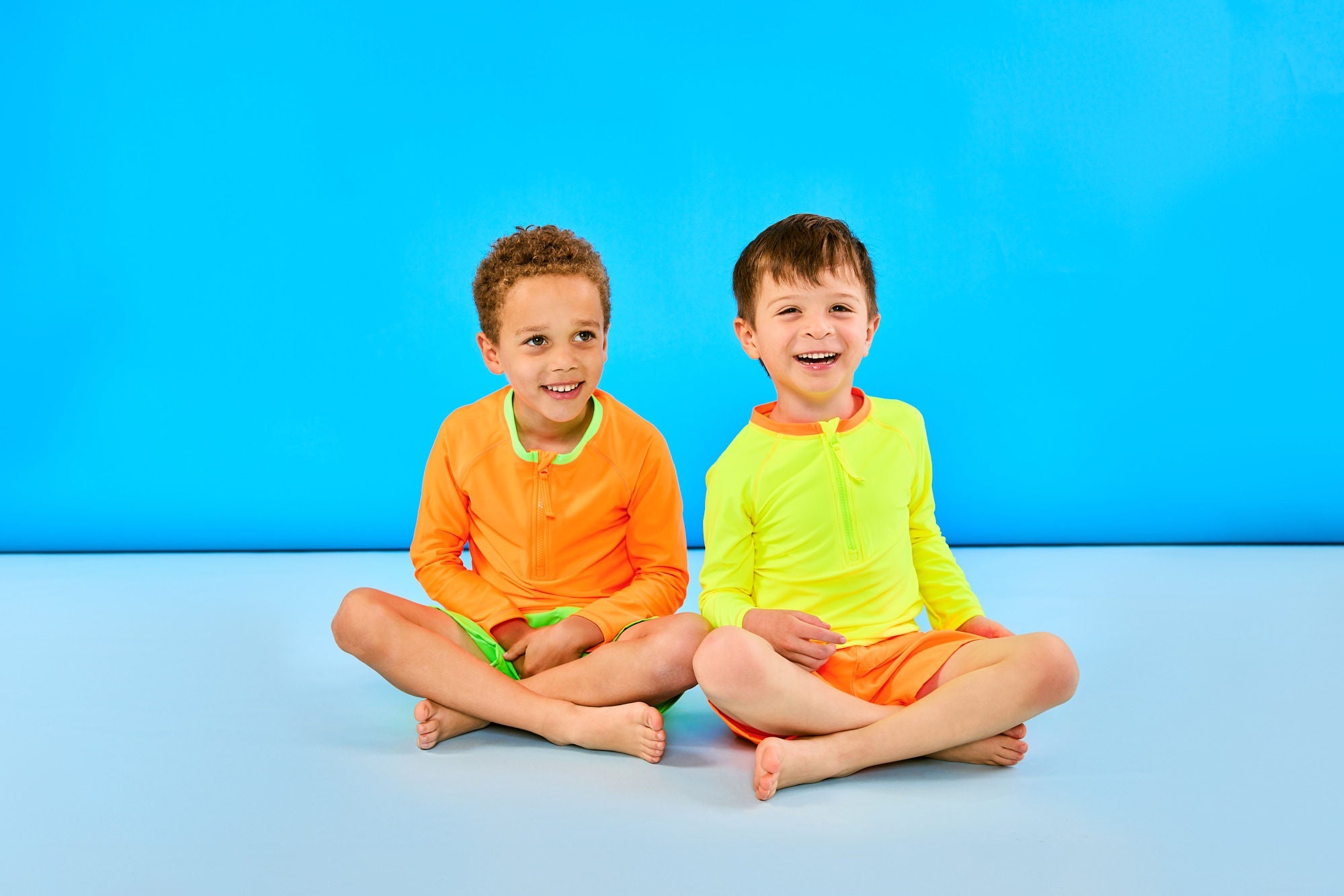 Two boys sitting. Boy on the left is smiling while wearing a neon orange rash guard and neon green swim trunks. The boy on the right is laughing while wearing a neon yellow rash guard and neon orange swim trunks.