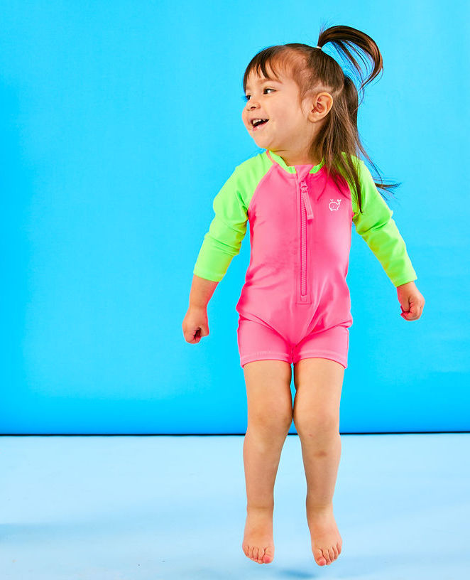 Toddler jumps playfully while wearing a neon pink and neon green rash guard one piece. The rash guard one piece features a zipper for easy on off and snaps for speedy diaper changes.