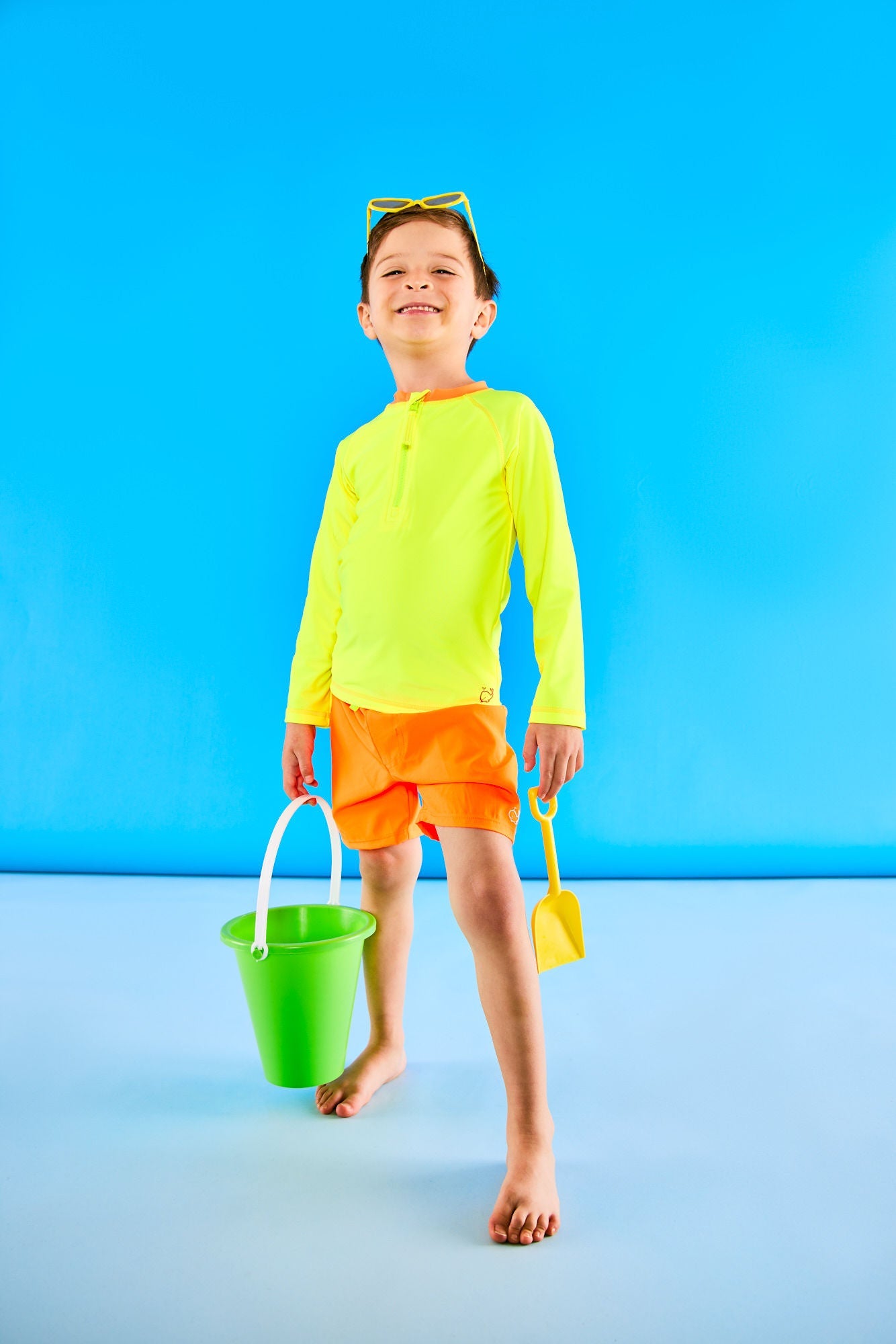 Young boy posing while wearing a neon yellow rashguard with a neon orange collar, neon orange swim trunks, and neon yellow sunglasses. The child is holding a green toy pail and a small yellow toy shovel