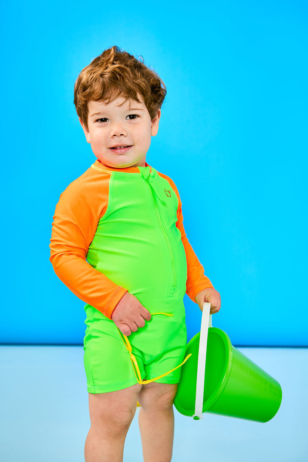 Toddler boy posing wearing a neon green and neon orange rashguard one piece. The toddler is holding sunglasses and a toy sand bucket.