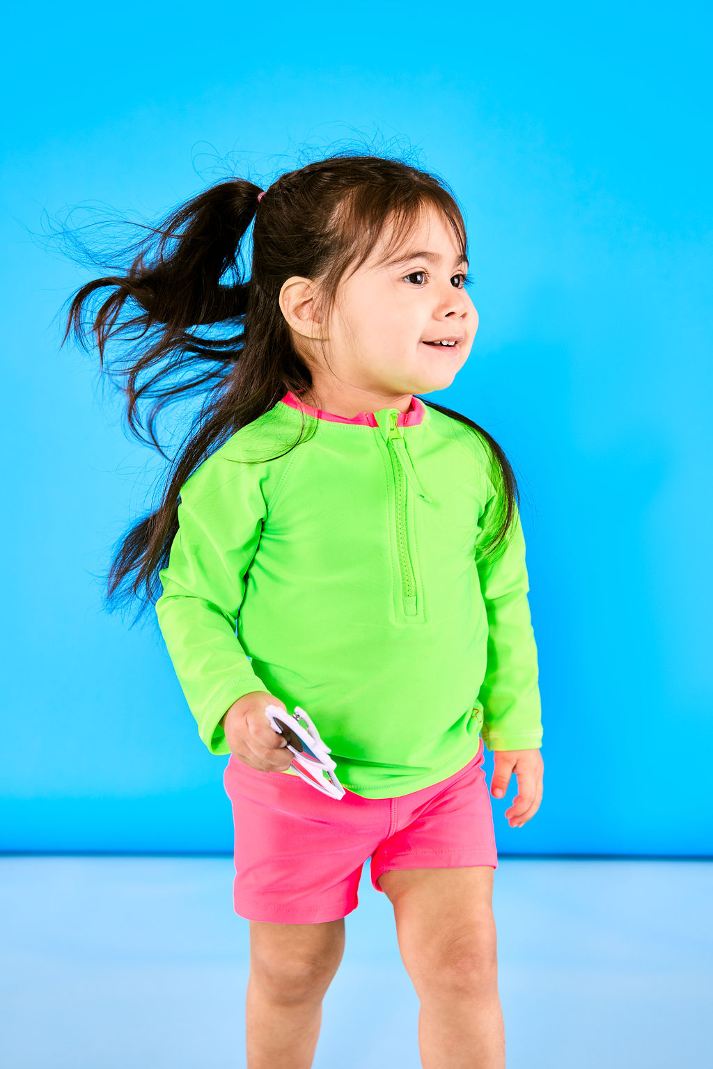 Toddler girl walking playfully wearing neon green rash guard and neon pink euro swim shorts while holding a pair of white sunglasses