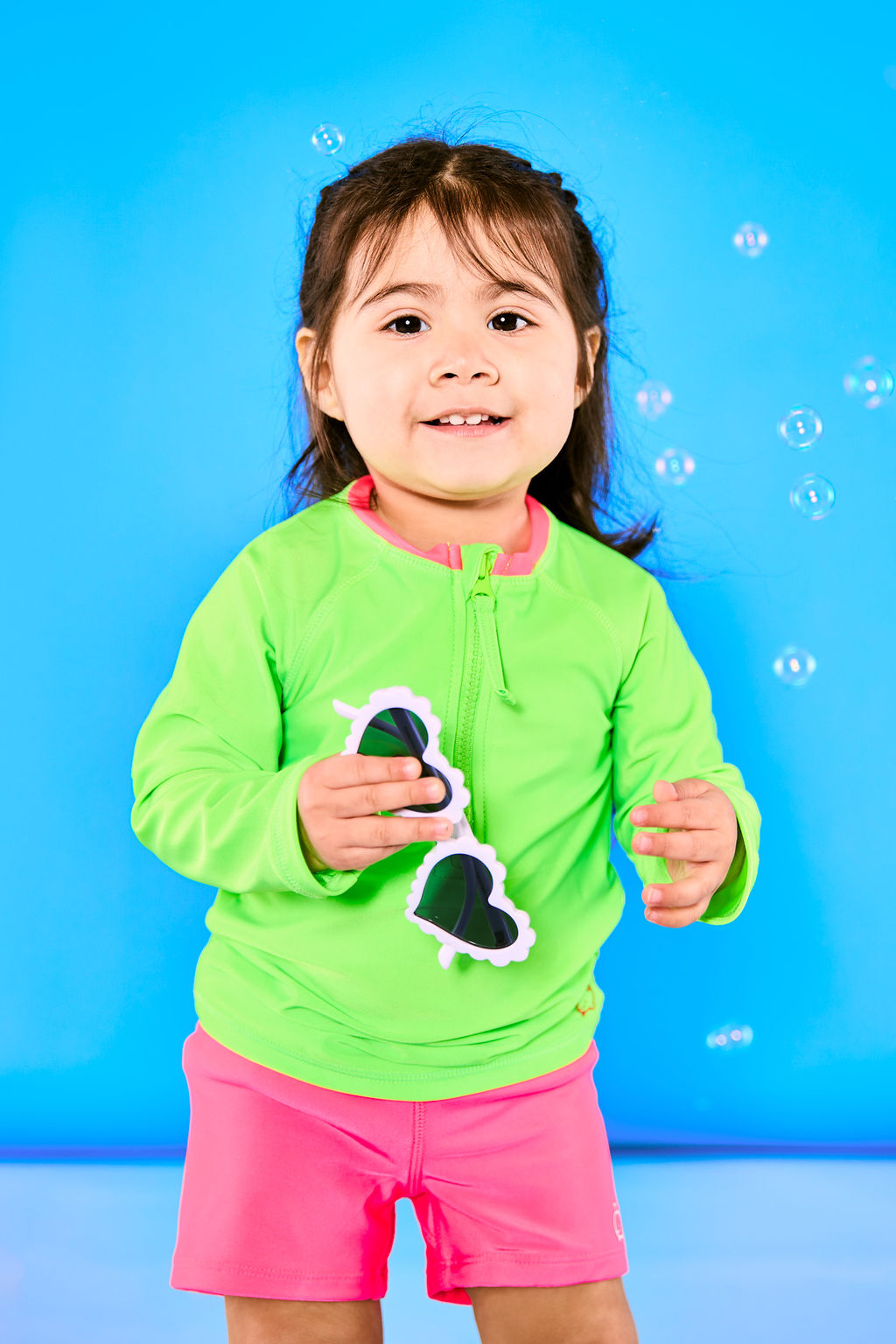 Child smiling and wearing neon green rash guard and neon pink euro swim shorts while holding a pair of white sunglasses and surrounded by bubbles.