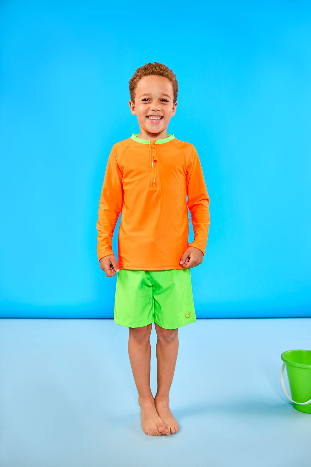 Child smiling and wearing children's neon orange rash guard with a neon green collar and neon green swim trunks.