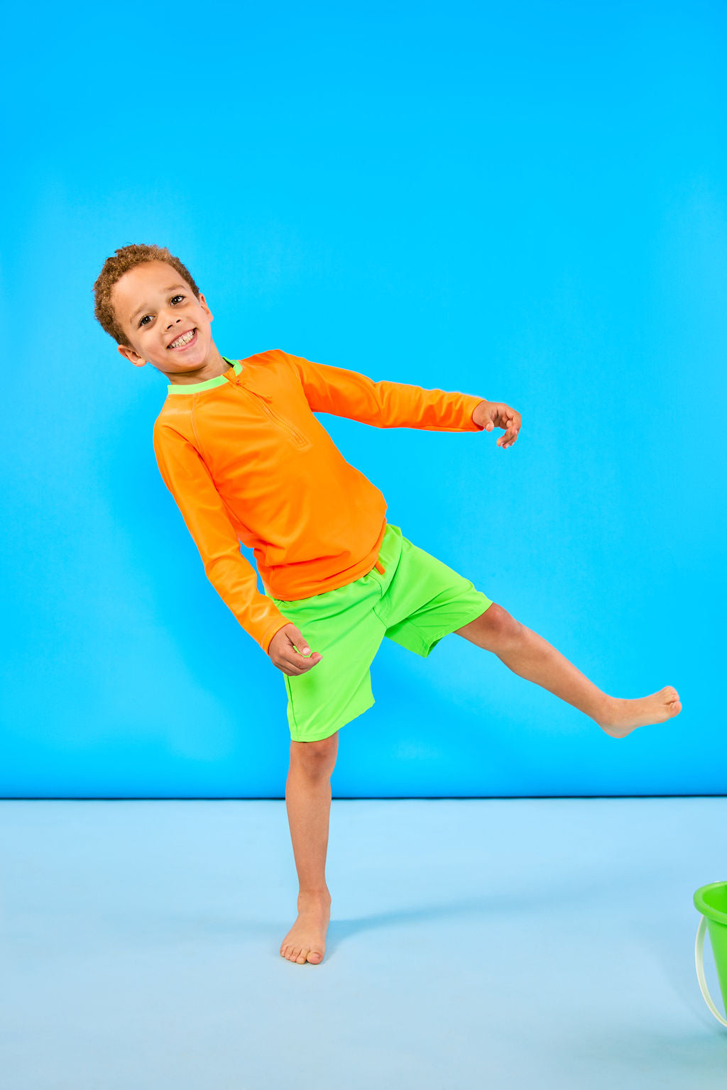 Child smiling and standing playfully while wearing children's neon orange rash guard with a neon green collar