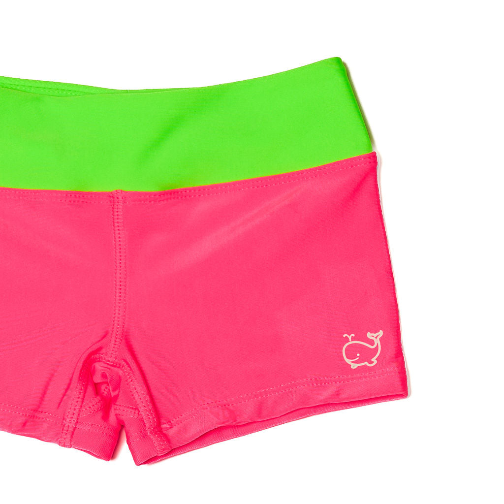 Close up of children's neon pink euro swim shorts with a neon green waistband. Shorts have a small white whale logo near the bottom hem.