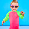 Toddler is posing playfully while wearing a neon pink and neon green rash guard one piece. The toddler is also wearing pink sunglasses.