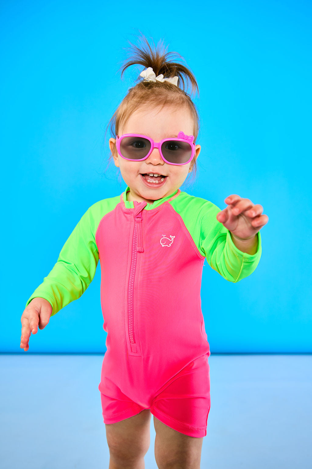 Toddler is posing playfully while wearing a neon pink and neon green rash guard one piece. The toddler is also wearing pink sunglasses.