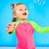 Toddler is playing with bubbles while wearing a neon pink and neon green rash guard one piece. The children's rash guard one piece features a zipper for easy on off.