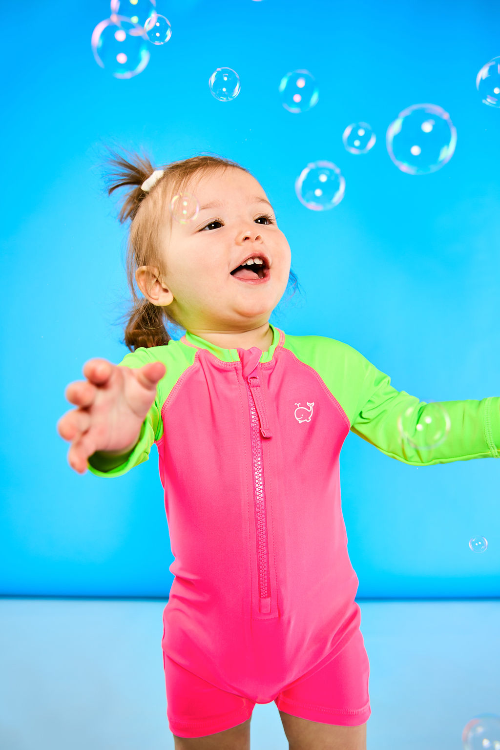 Toddler is playing with bubbles while wearing a neon pink and neon green rash guard one piece. The children's rash guard one piece features a zipper for easy on off.