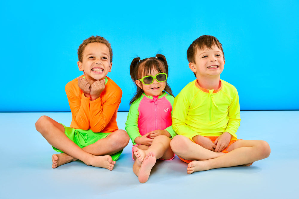 Three children smiling wearing neon swimsuits. Child on the left looks excited wearing neon orange shirt and neon green shorts. Middle child is smiling wearing neon green sunglasses and a neon pink and green swimsuit. Child on the right is smiling wearing neon yellow and neon orange.