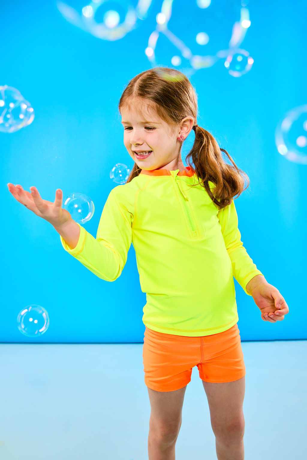 Child playing with bubbles while wearing a neon yellow and neon orange rash guard that features a zipper for easy on off. The child is also wearing neon orange euro swim shorts with a neon yellow waistband.