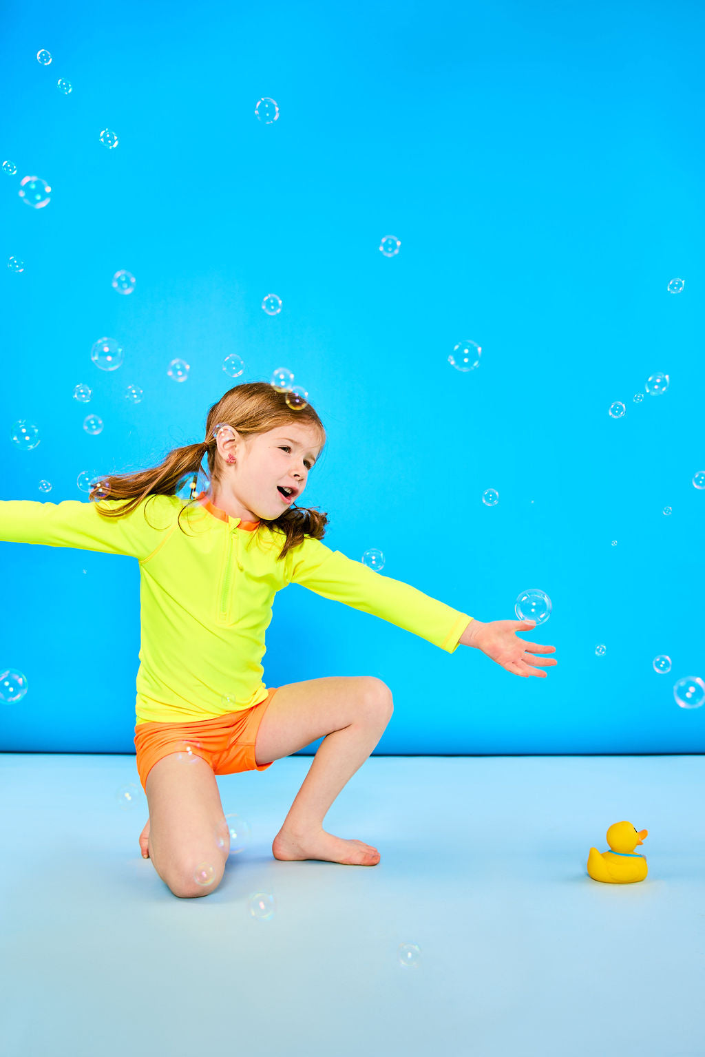 Child posing playfully wearing a neon yellow rash guard and neon orange euro swim shorts while surrounded by bubbles and next to a small rubber duck