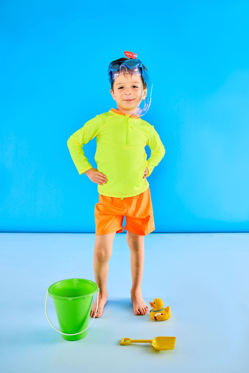 Child posing playfully with beach toys while wearing neon yellow sunglasses and neon yellow rash guard and neon orange swim trunks.