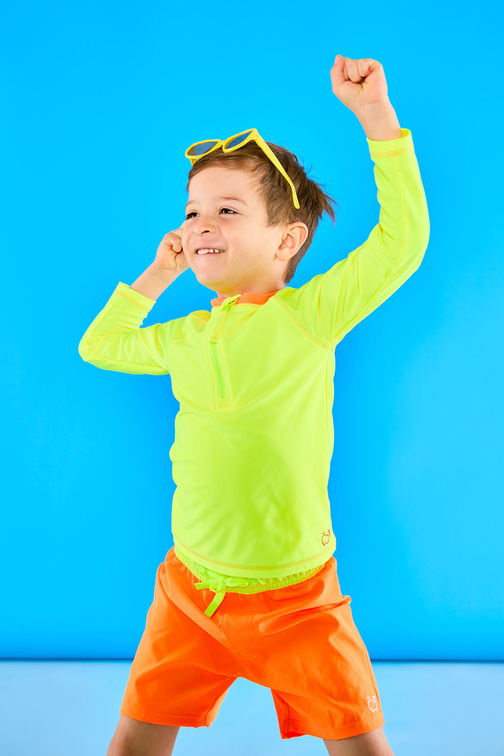 Child posing playfully wearing neon yellow sunglasses and neon yellow rash guard and neon orange swim trunks